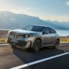 BMW iX4 50 xDrive, SUV coupé électrique gris-vert, en roulage sur une route côtière face à la mer et aux montagnes (photo officielle BMW)