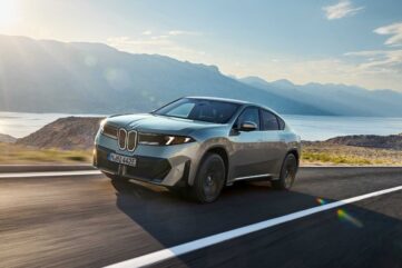 BMW iX4 50 xDrive, SUV coupé électrique gris-vert, en roulage sur une route côtière face à la mer et aux montagnes (photo officielle BMW)
