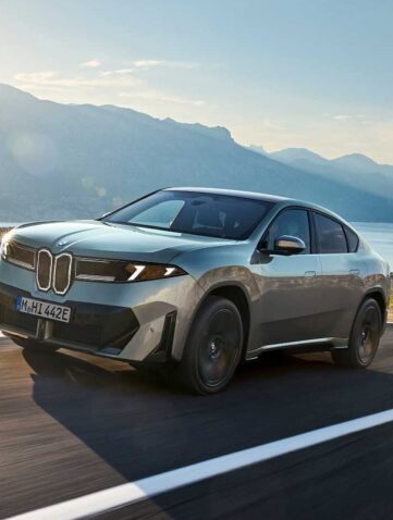 BMW iX4 50 xDrive, SUV coupé électrique gris-vert, en roulage sur une route côtière face à la mer et aux montagnes (photo officielle BMW)