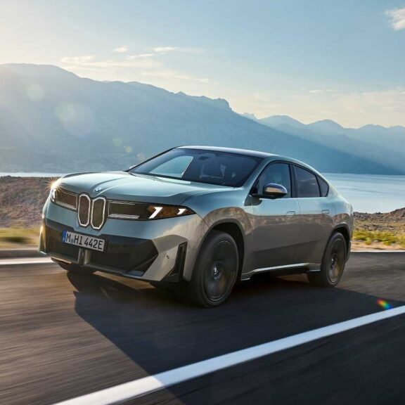 BMW iX4 50 xDrive, SUV coupé électrique gris-vert, en roulage sur une route côtière face à la mer et aux montagnes (photo officielle BMW)